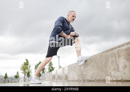 Niedrigwinkelaufnahme von fittem älteren Mann in Sportbekleidung dehnend Beinmuskeln in Ausfallposition auf Betonstufen gegen bewölkten Himmel im Stadtpark, Kopierraum Stockfoto