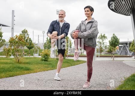 Eine vollständige Aufnahme von lächelnden älteren Männern und Frauen, die Knie umarmen, sich aufwärmen und sich auf das Joggen im morgendlichen Stadtpark mit grauem Himmel vorbereiten Stockfoto