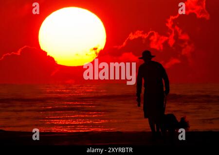 Isle Of Palms, Usa. 30. August 2024. Ein Mann spaziert mit seinem Hund entlang des von der Sonne umrahmten Strandes, während er über dem Atlantischen Ozean in Wild Dunes, 30. August 2024 in Isle of Palms, South Carolina, aufgeht. Quelle: Richard Ellis/Richard Ellis/Alamy Live News Stockfoto