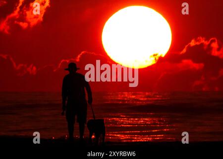 Isle Of Palms, Usa. 30. August 2024. Ein Mann spaziert mit seinem Hund entlang des von der Sonne umrahmten Strandes, während er über dem Atlantischen Ozean in Wild Dunes, 30. August 2024 in Isle of Palms, South Carolina, aufgeht. Quelle: Richard Ellis/Richard Ellis/Alamy Live News Stockfoto