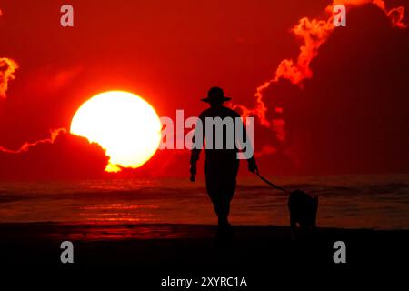 Isle Of Palms, Usa. 30. August 2024. Ein Mann spaziert mit seinem Hund entlang des von der Sonne umrahmten Strandes, während er über dem Atlantischen Ozean in Wild Dunes, 30. August 2024 in Isle of Palms, South Carolina, aufgeht. Quelle: Richard Ellis/Richard Ellis/Alamy Live News Stockfoto