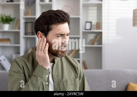 Mann mit Ohrenschmerzen, der auf dem Sofa in der häuslichen Umgebung sitzt. Der Ausdruck von Unwohlsein deutet auf mögliche Ohrenschmerzen oder Entzündungen hin. Casual verbessert das Gefühl des persönlichen Kampfes mit Gesundheitsproblemen. Stockfoto