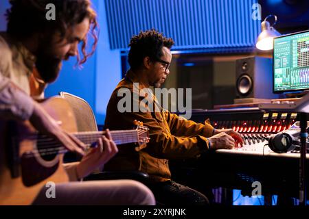 Künstler spielt ein Lied auf elektroakustischer Gitarre im professionellen Studio, nimmt Tracks auf Musikinstrumenten auf, um mit seinem Techniker neue Musik zu produzieren. Sänger komponiert Schallplatten im Kontrollraum. Stockfoto