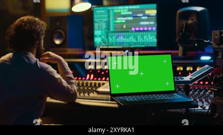 Audiotechniker, der den Ton auf der Mischkonsole des Recorders ausgleicht, mit einem Laptop, auf dem die Greenscreen-Anzeige angezeigt wird. Der Produzent dreht die Lautstärke der Titel im professionellen Studio, im Kontrollraum. Kamera A.. Stockfoto