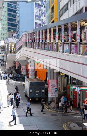 Hongkong, China - 03. Juli 2024 : Eine belebte Straße in Hongkong mit Fußgängern, die auf einem Gehsteig aus Beton unter einem Fußgängerweg laufen. Stockfoto