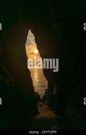 Eine Höhle mit einer kleinen Öffnung und der Sonne, die durch sie hindurchscheint. Die Sonne untergeht und das Wasser ist ruhig. Stockfoto