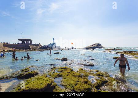 Kanyakumari, Indien, 22. Februar 2015: Indische Touristen schwimmen am flachen felsigen Strand mit Thiruvalluvar-Statue und 16-beinigem Mandapfelpavillon hinten Stockfoto