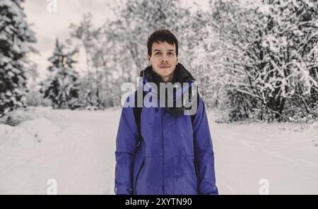 Junger erwachsener Mann mit braunen, kurzen Haaren, der während des Winters in norwegen in einer wunderschönen Schneelandschaft steht, eine blaue Regenjacke trägt, die Kamera anschaut, NOR Stockfoto