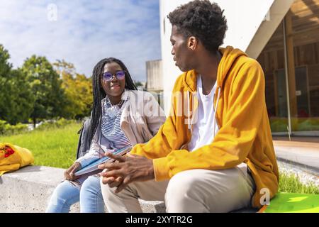 Seitenansicht zweier männlicher und weiblicher afrikanischer Studenten, die sich entspannt auf dem Campus unterhalten und auf einer Bank sitzen Stockfoto