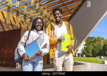 Zwei junge junge, selbstbewusste afroamerikanische Studenten, die zusammen auf dem Universitätsgelände laufen, tragen Schultasche und -Ordner Stockfoto