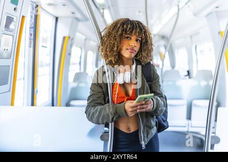 Lateinische Frau mit lockigem Haar, die im Zug mit Handy steht und in die Kamera schaut Stockfoto