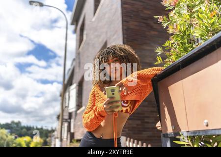 Selbstbewusste lateinfrau, die ein Telefon benutzt, das sich in einer Fassade in einem Wohnviertel unter der Sonne lehnt Stockfoto