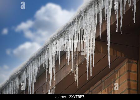 Kristalline und glitzernde Eiszapfen, die sich aufgrund der eisigen Witterungsbedingungen gebildet haben und am Rand eines braunen hölzernen Wohnhauses hängen Stockfoto