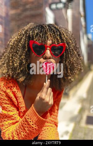 Vertikales Foto einer lateinischen verspielten Frau mit Sonnenbrille, die einen Herzlutscher auf der Straße hält Stockfoto