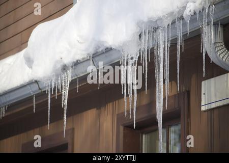 Kristalline und glitzernde Eiszapfen, die sich aufgrund der eisigen Witterungsbedingungen gebildet haben und am Rand eines braunen hölzernen Wohnhauses hängen Stockfoto