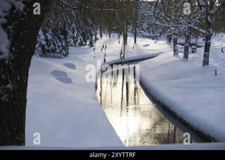 Ruhige Winterszene mit einem kleinen, ruhigen Bach, der durch einen schneebedeckten Park mit den frostbeladenen Ufern und Bäumen, die ihn umgeben, Bäumen und dem Stockfoto