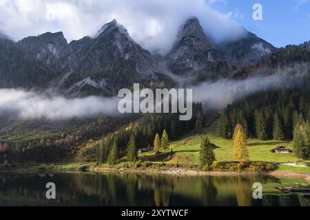 Der vordere Gosausee im Herbst mit Blick auf den wolkenbedeckten Gosaukamm. Die Seeklausalm in der Mitte zwischen den Bäumen. Bewölkter und blauer Himmel mit s Stockfoto