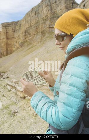 Ein Reisender mit Hut und Sonnenbrille hält hundert Dollar-Scheine in den Händen eines Ventilators vor dem Hintergrund von Felsen auf der Natur. Reisekosten Stockfoto