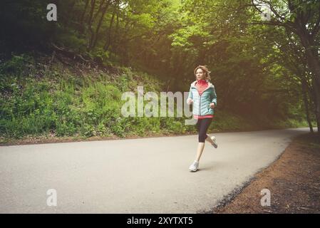 Junge Fitness blonde Frau in Kopfhörer läuft am Morgen kaukasischen Wald Trail in Sonnenlicht Stockfoto