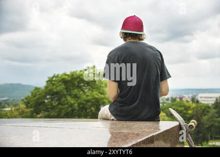 Der langhaarige Skater in Kappe und T-Shirt sitzt mit dem Rücken zur Kamera und denkt neben dem Skateboard vor dem Hintergrund der Landschaft Stockfoto