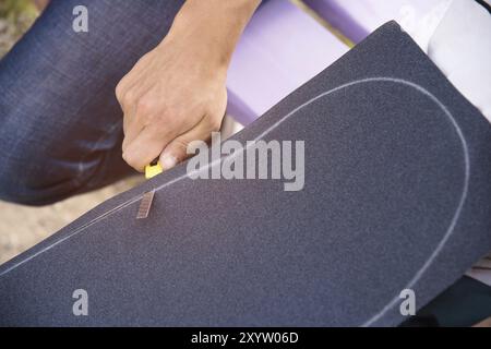 Ein kleiner Junge auf den Knien klebt das Griptape in Begleitung von Freunden bei sonnigem Wetter auf ein Skateboard. Vorbereitung eines Skateboards für einen Skatepark-Wettkampf Stockfoto