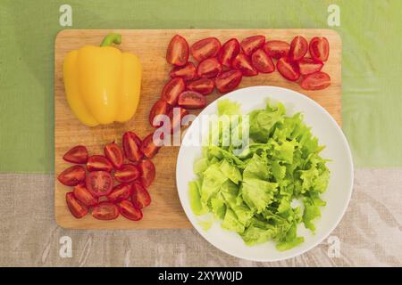 Frisch gehackte grüne Salatblätter liegen auf einem Holzschneidebrett neben der gelben Paprika und gehackten roten Kirschtomaten auf einem grünen Tisch. Die Stockfoto