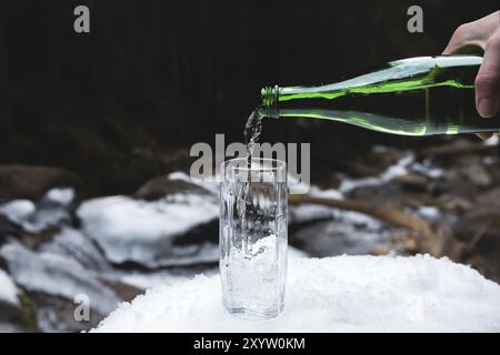 Mineralwasser wird aus einer glasgrünen Flasche in ein klares Glas-Becherglas gegossen. Ein Glas steht im Schnee. Vor dem Hintergrund eines Winters Stockfoto