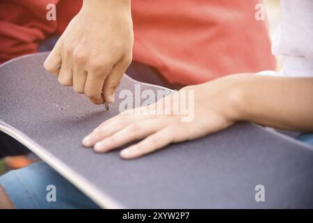 Ein kleiner Junge auf den Knien klebt das Griptape in Begleitung von Freunden bei sonnigem Wetter auf ein Skateboard. Vorbereitung eines Skateboards für einen Skatepark-Wettkampf Stockfoto