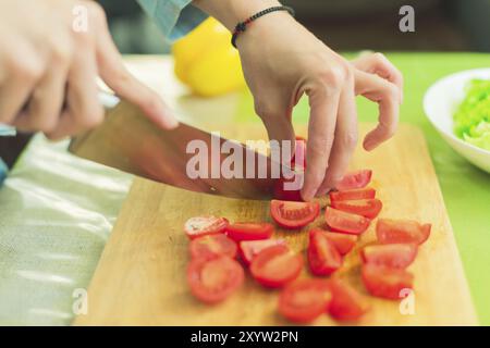 Die Hände eines jungen Mädchens hacken die Kirschtomaten auf einem hölzernen Schneidebrett auf einem grünen Tisch in einem Wohnbereich gegen einen Teller mit geschnittenen Salatblättern Stockfoto