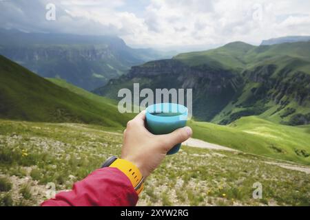 Aus erster Person betrachtet, wie die Hand eines Mannes eine Tasse Tee aus Plastik gegen ein Plateau aus grünen Hügeln hält und im Sommer einen wolkenlosen Himmel hat Stockfoto