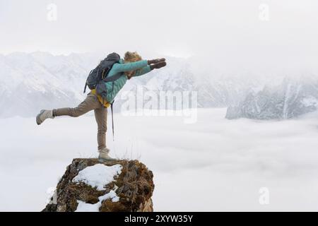 Ein fröhlich lächelndes Hipster-Mädchen reist in einer Daunenjacke mit Rucksack und in einem großen Pelzmütze steht wieder in einer superman-Flugposition auf einem Felsen Stockfoto