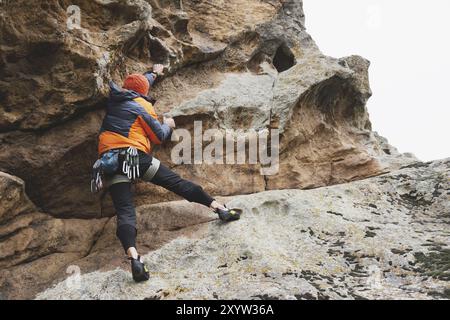 Hipster, Kletterer im Alter, in dem man einen wunderschönen Felsen ohne Versicherung und Helm hochklettert. Ein Bergsteiger in Hut und Daunenjacke mit Tasche für magnes Stockfoto