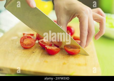 Die Hände eines jungen Mädchens hacken die Kirschtomaten auf einem hölzernen Schneidebrett auf einem grünen Tisch in einem Zuhause. Das Konzept von gesunder Ernährung und Vegetarisch Stockfoto