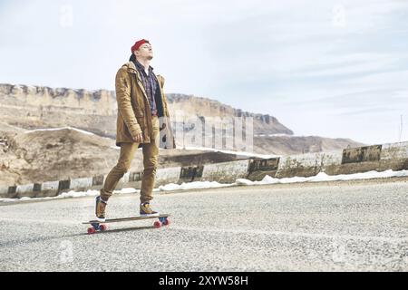 Stylischer Happy Young man in einer Mütze und Hose Jogginghose, die auf einem Longboard eine Bergstraße hinunterrollt und das Leben genießt Stockfoto