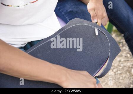 Ein kleiner Junge auf den Knien klebt das Griptape in Begleitung von Freunden bei sonnigem Wetter auf ein Skateboard. Vorbereitung eines Skateboards für einen Skatepark-Wettkampf Stockfoto