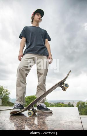 Ein junger langhaariger Skater in T-Shirt und durchgehender Cap steht mit einem Skateboard vor dem Hintergrund der Stadt Stockfoto