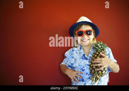 Ein hübsches Kind trägt einen Hut und eine Sonnenbrille, die Ananas auf rotem Hintergrund hält. Kopierraum, Tageslicht Stockfoto