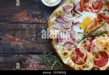 Pizza Carbonara mit Speck, Salami, Parmesan-Käse und Huhn-Ei am rustikalen Holztisch Stockfoto