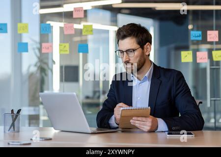 Business Professional in Anzug arbeitet fleißig am Laptop, überprüft Daten und macht Notizen am Schreibtisch. Die Umgebung umfasst farbenfrohe Haftnotizen auf Glas, die kreative Ideen widerspiegeln. Stockfoto