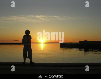 Kleine Jungen fischen bei Sonnenuntergang auf einem Pier im Bagenkop Hafen auf der dänischen Insel Langeland. Kleiner Junge fischt bei Sonnenuntergang auf einem Pier in Bage Stockfoto