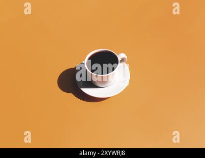 Eine weiße Tasse und Unterteller mit schwarzem Espresso-Kaffee von oben mit Schatten der Sonne auf gelbem Terrakotta-Hintergrund. Stockfoto