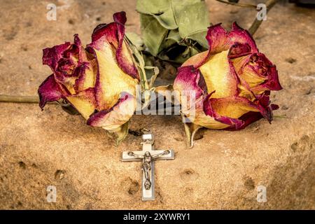Christliches Kreuz auf einem Rosenkranz und eine trockene gelb-rote Rose, die auf einem roten Sandstein liegt Stockfoto