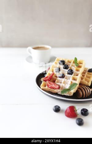 Stapel Waffeln auf einem Teller auf dem weißen Tisch mit Blaubeeren, gehackten Erdbeeren- und Minzblättern und einer weißen Tasse Kaffee. Stockfoto