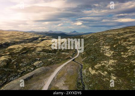 Die gewundene Straße schlängelt sich durch die zerklüftete Landschaft des Rondane-Nationalparks in Norwegen, bedeckt mit Flechten und Heidekraut unter einem hellen, sonnendurchfluteten Himmel Stockfoto