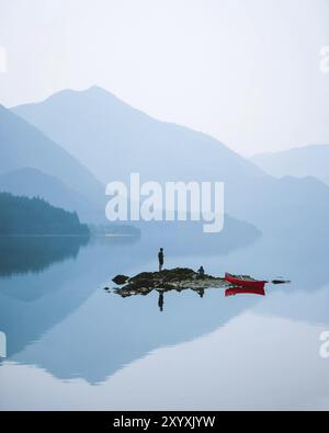 Person auf einer kleinen Insel in einem nebeligen See, umgeben von Bergen, mit einem roten Kanu in der Nähe, Slocan Lake, British Columbia, Kanada, North Ameri Stockfoto