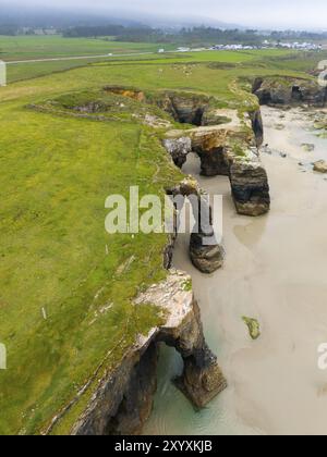 Steile Klippen mit mehreren markanten Felsbögen und saftig grünen Wiesen, die bis zum Meer reichen, aus der Vogelperspektive, Praia das Catedrais, Playa de las Catedr Stockfoto