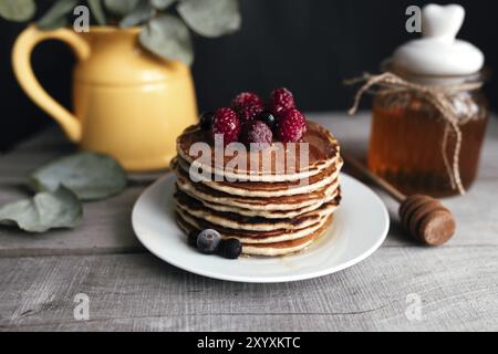 Saftige Pfannkuchen mit Beeren und Honig auf einem weißen Teller, Löffel, Glas, Holztisch, gelbe Vase mit Eukalyptus. Stockfoto