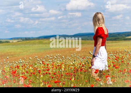 Blonde junge Frau, die in einem rot-weißen Kleid auf einem Feld mit rotem Mohn steht Stockfoto