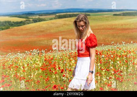 Blonde junge Frau, die in einem rot-weißen Kleid auf einem Feld mit rotem Mohn steht Stockfoto