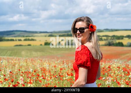 Blonde junge Frau in Sonnenbrille, rotes weißes Kleid in einem Feld aus rotem Mohn mit rotem Mohn im Haar Stockfoto